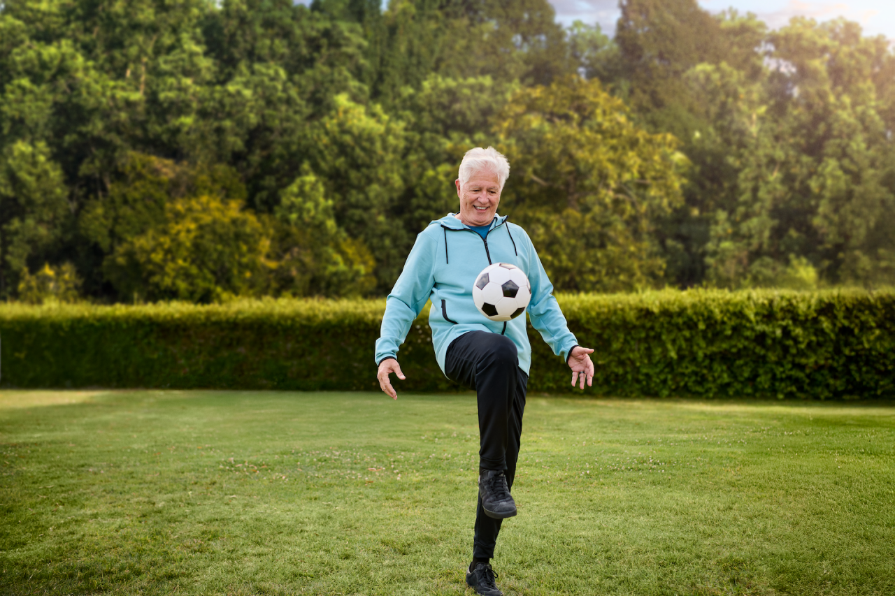 Man with white hair and blue jacket, using his feet to juggle a soccer ball in green grass